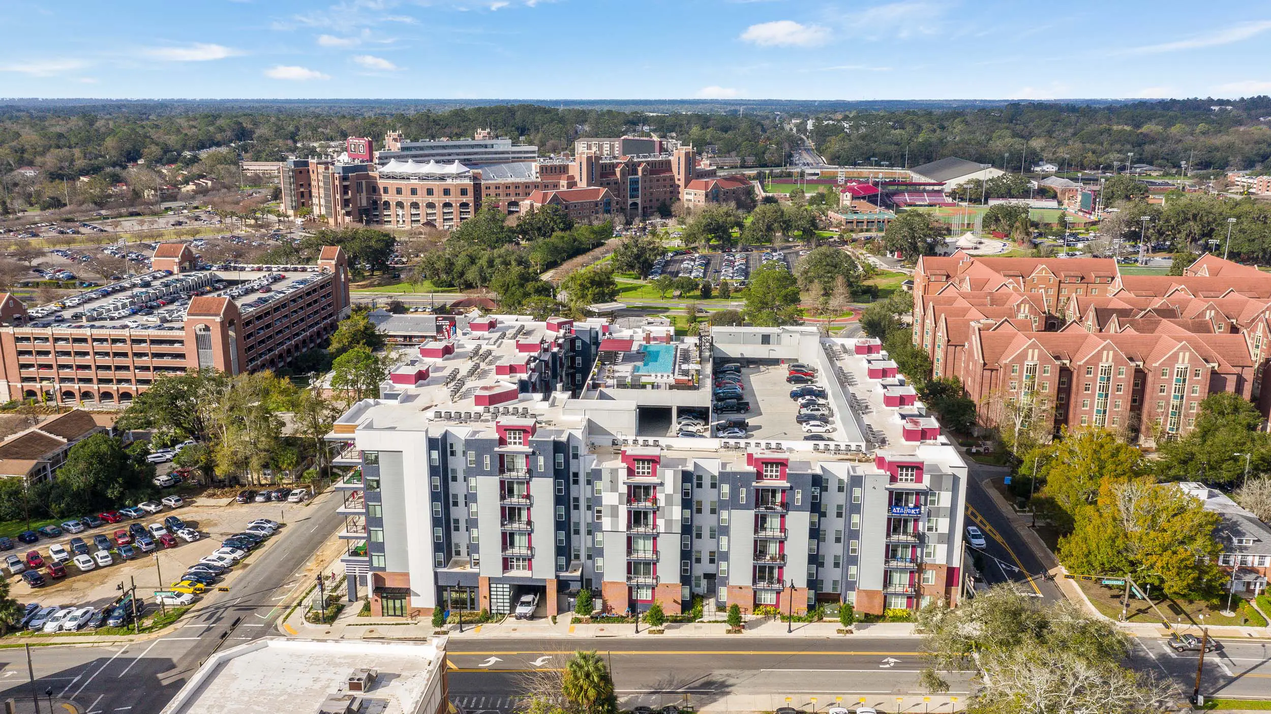 Drone view of a six-story student-housing building showing the rooftop pool and part of the parking deck.