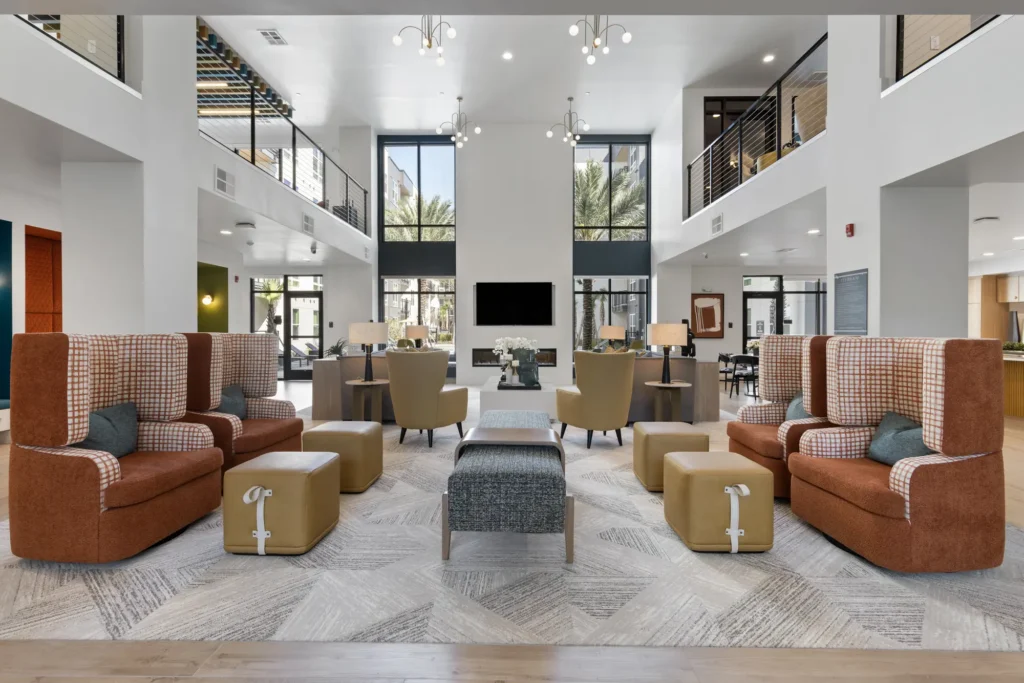 View from the back of a two-story common area at the Aston at Uptown with couches, chairs, ottomans, and recessed seating nooks. A TV is mounted above a gas fireplace.