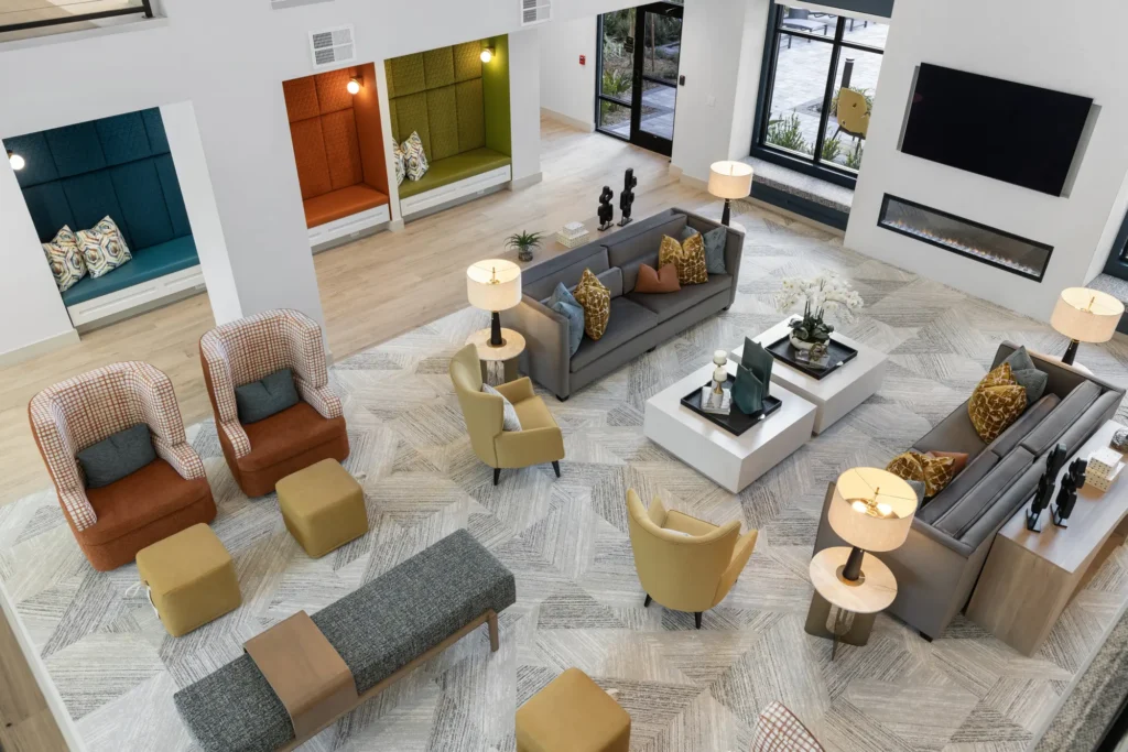 View from above looking down into a two-story common area at the Aston at Uptown with couches, chairs, ottomans, and recessed seating nooks. A TV is mounted above a gas fireplace.