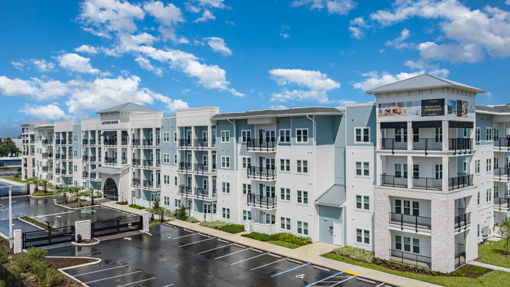 Four-story Lantower Bayside facade at twilight, alternating white stucco and blue-grey siding, warm-lit windows and black-railed balconies above a lawn and walkway