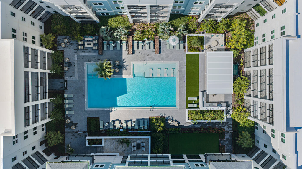 Top-down view of the pool courtyard: a turquoise pool with in-water loungers, grey-tiled patios with lounge and dining furniture, artificial turf, and white balconied buildings enclosing both sides