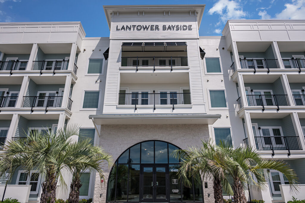 The arched glass entry of Lantower Bayside framed by stone-textured walls and two palms, with columns of black-railed balconies stacked above