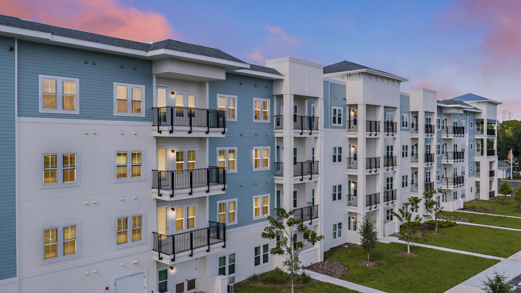 Street-level view of Lantower Bayside's exterior walk: stacked black-railed balconies on white piers, blue-grey siding and a white stone accent wall behind shrubs and ornamental grasses